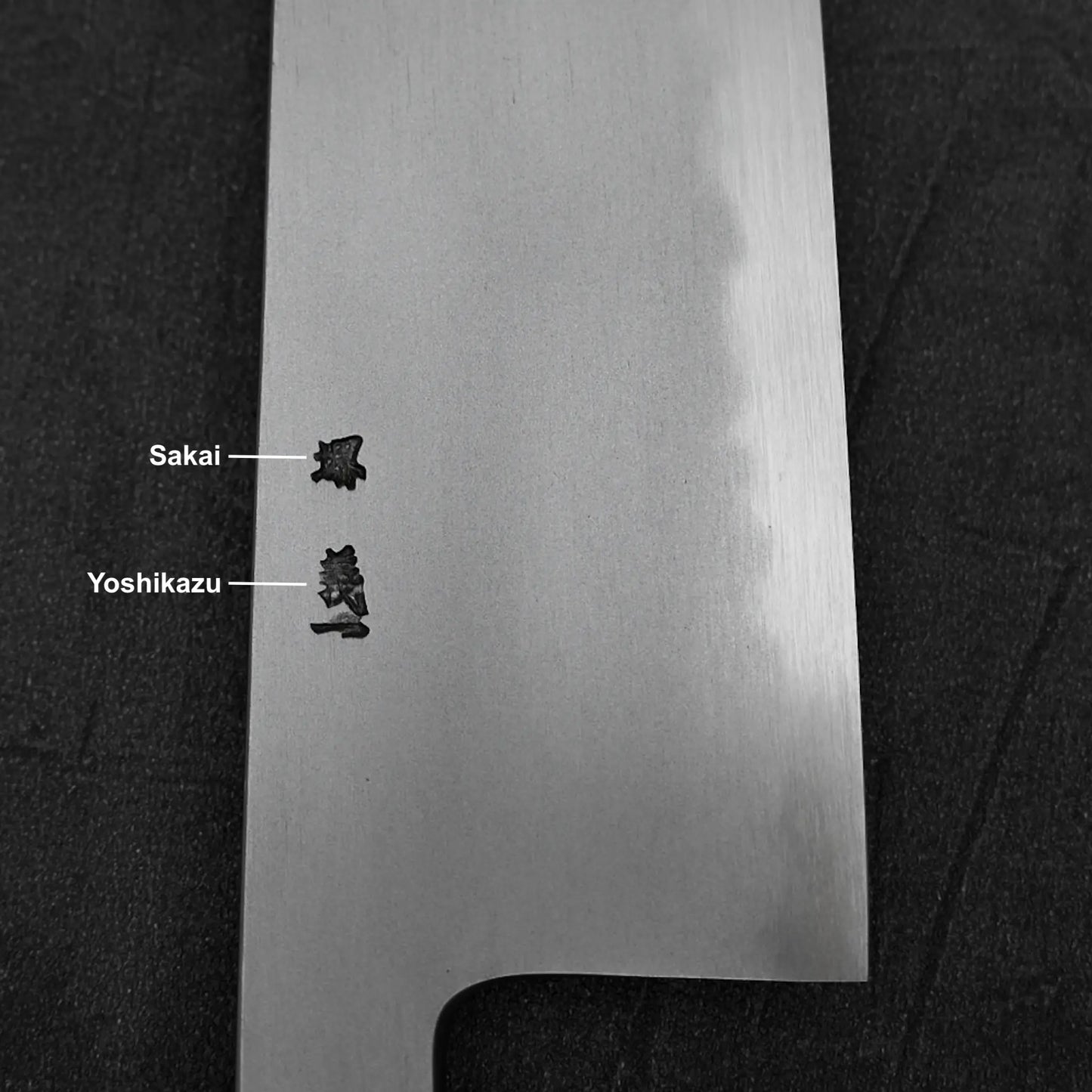 A close-up of the Yoshikazu Tanaka AS tall gyuto 240mm blade shows two kanji engravings: Sakai for the renowned Sakai tradition and Yoshikazu for master blacksmith Yoshikazu Tanaka. The knife rests on a black surface.