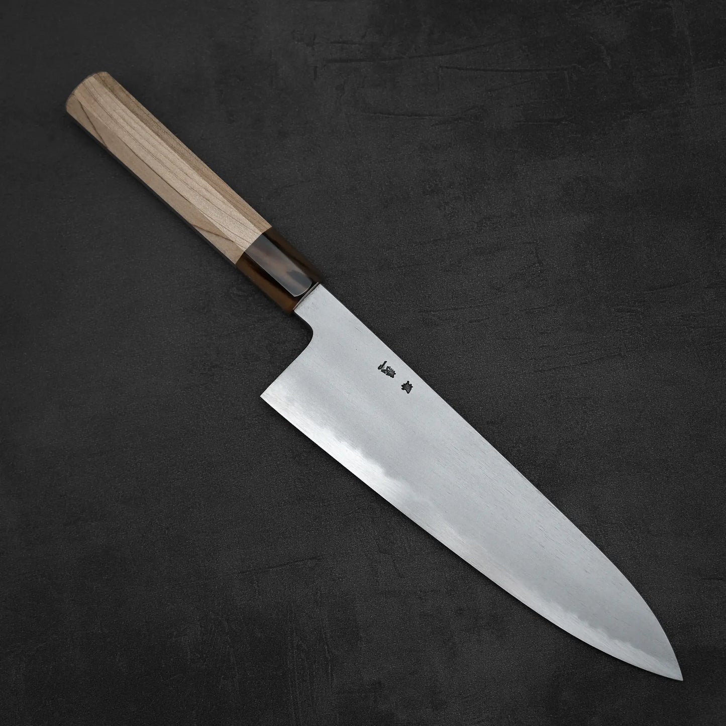The Yoshikazu Tanaka AS tall gyuto 240mm, featuring a light wooden handle and stainless steel blade with subtle Japanese characters near the handle, rests on a dark, textured surface.