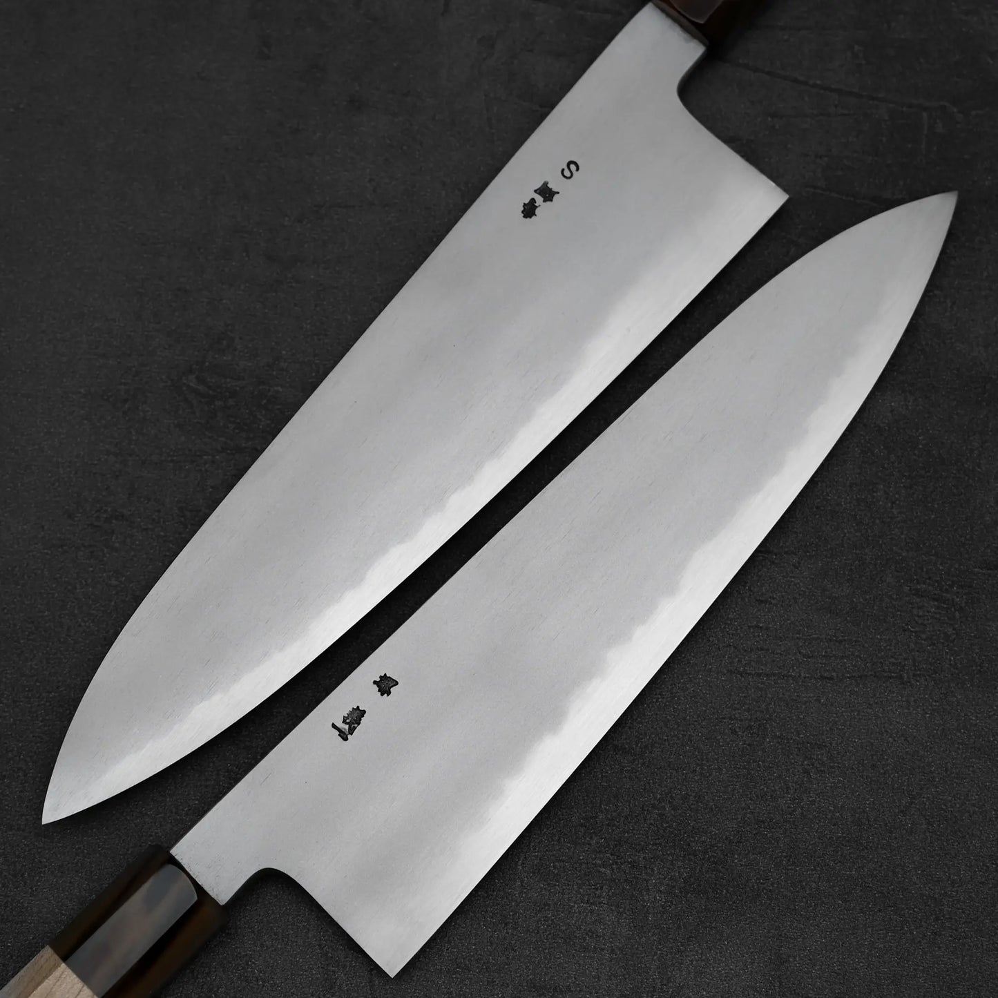 Two Yoshikazu Tanaka AS tall gyuto 240mm chef’s knives, crafted by the renowned Sakai blacksmith, feature wooden handles and Japanese characters. Their polished blades with sharp edges are set diagonally on a dark, textured surface.