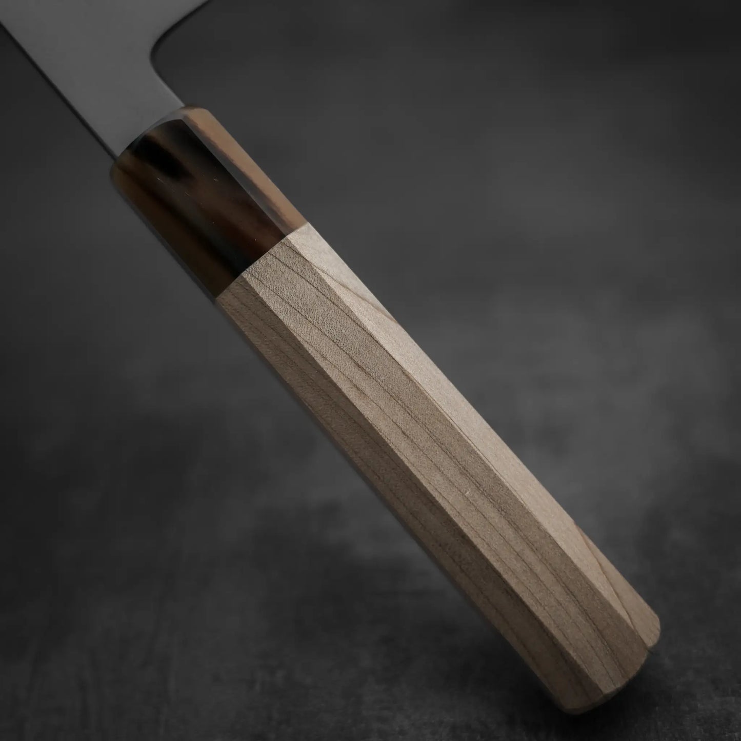 A close-up of the Yoshikazu Tanaka AS tall gyuto 240mm shows its light wood handle with a darker, glossy section near the blade, set against a dark, blurred background—highlighting the craftsmanship of Yoshikazu Tanaka.