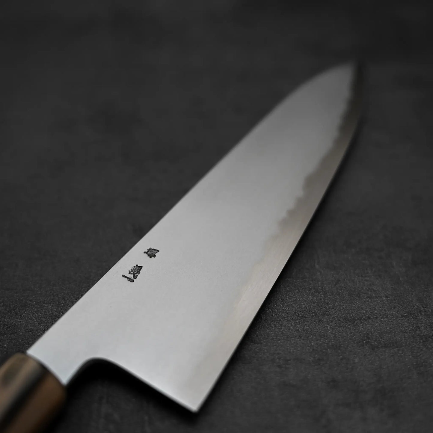 A close-up of the Yoshikazu Tanaka AS tall gyuto 240mm chefs knife, featuring a wooden handle and delicate blade engravings, from renowned blacksmith Yoshikazu Tanaka, displayed on a dark gray textured background.