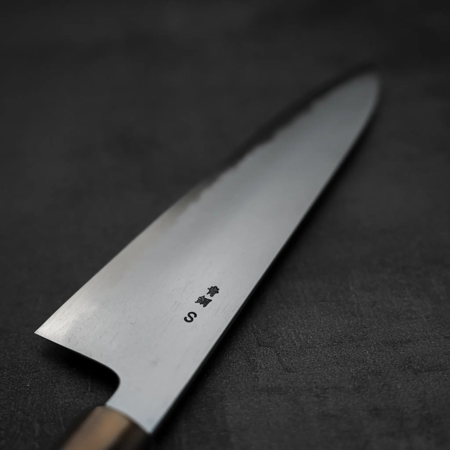 A close-up of the Yoshikazu Tanaka AS tall gyuto 240mm chefs knife by Yoshikazu Tanaka, featuring Japanese characters and an S engraved on the blade, shown on a dark textured surface.