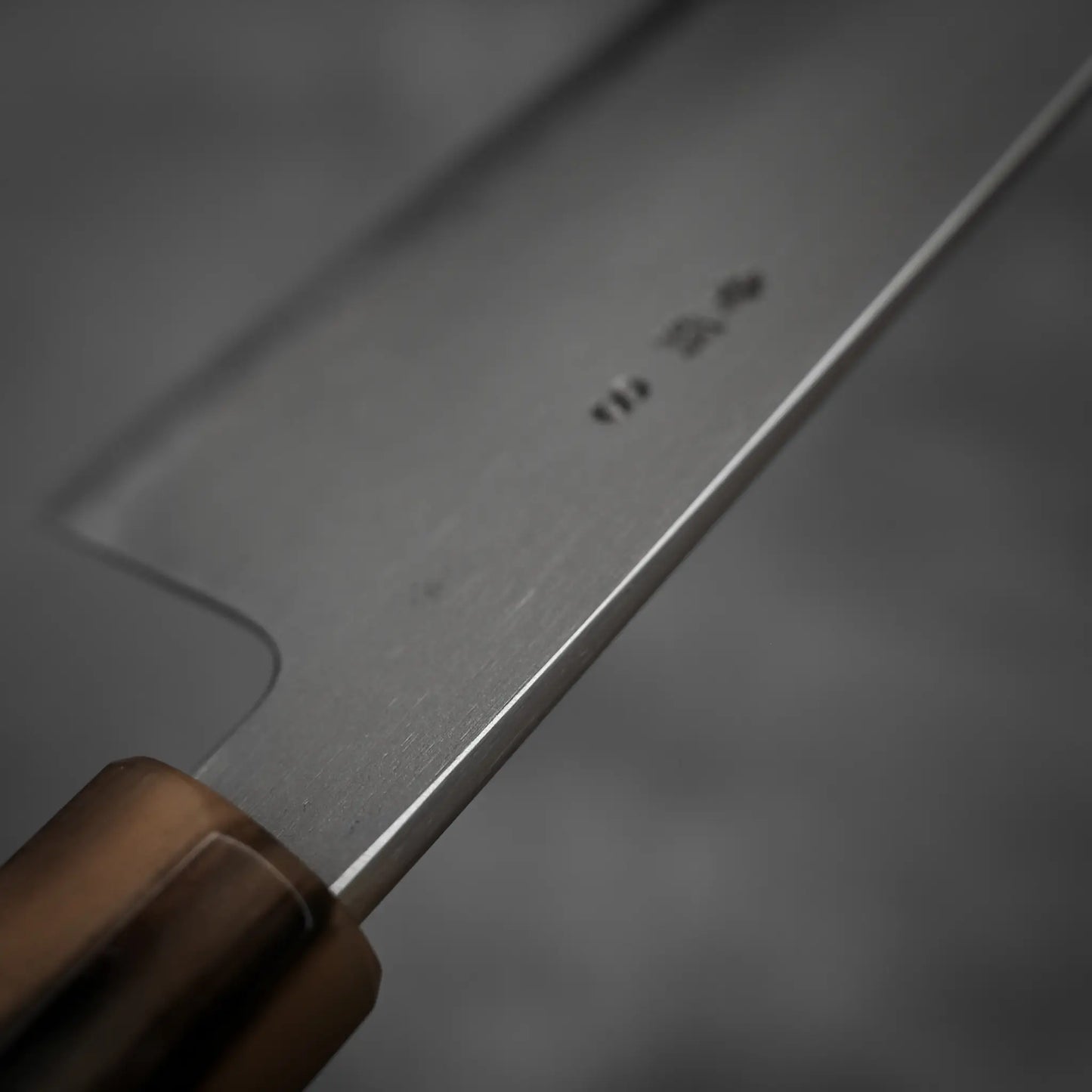 Close-up of the Yoshikazu Tanaka AS tall gyuto 240mm with a wooden handle and sharp stainless steel blade, faint markings visible, photographed against a blurred gray background.