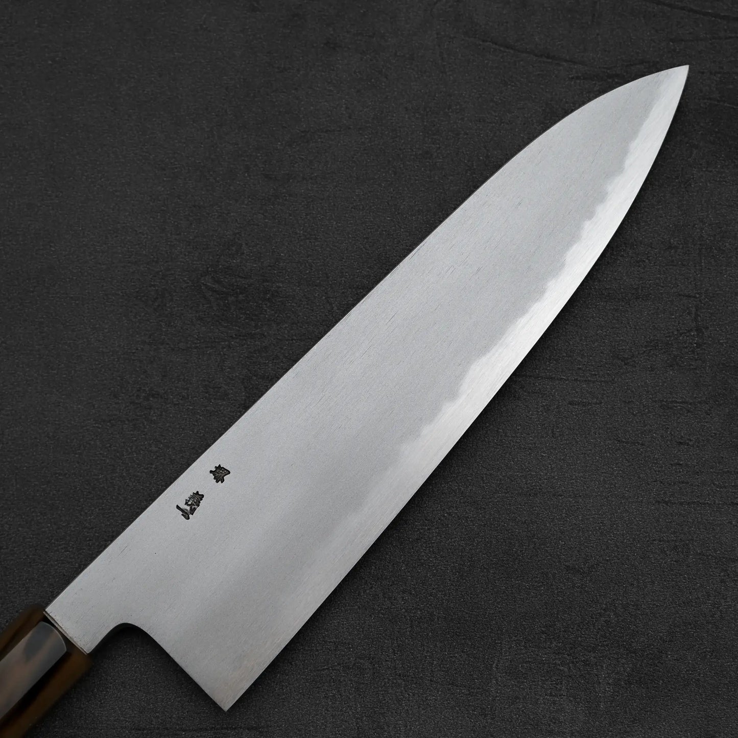 A close-up of the Yoshikazu Tanaka AS tall gyuto 240mm, crafted by Sakai blacksmiths, lies on a dark surface. The blade displays a sharp edge and engraved characters near the handle, showcasing Yoshikazu Tanakas artistry.