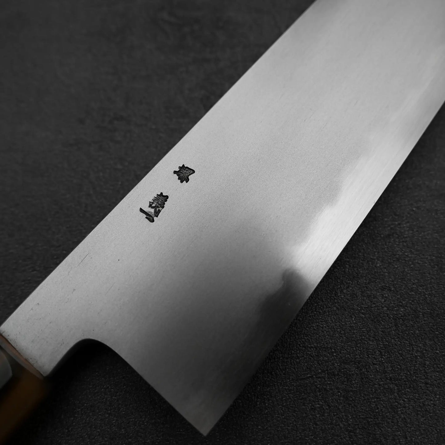 A close-up of the Yoshikazu Tanaka AS tall gyuto 240mm knife, featuring a wooden handle and engraved kanji on the blade, by Yoshikazu Tanaka, rests on a dark textured surface.