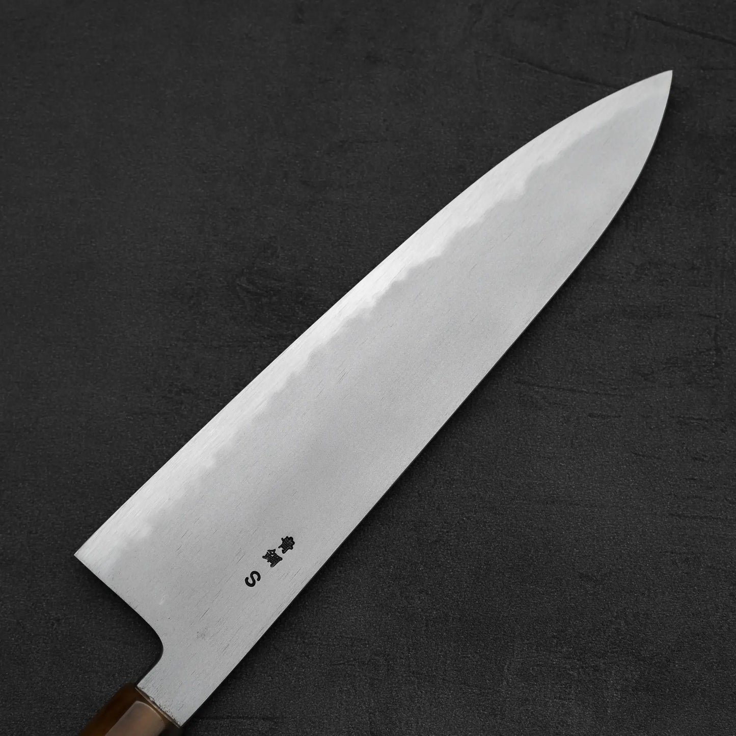 A close-up of the Yoshikazu Tanaka AS tall gyuto 240mm knife shows its wooden handle and polished, engraved blade resting on a dark textured surface.