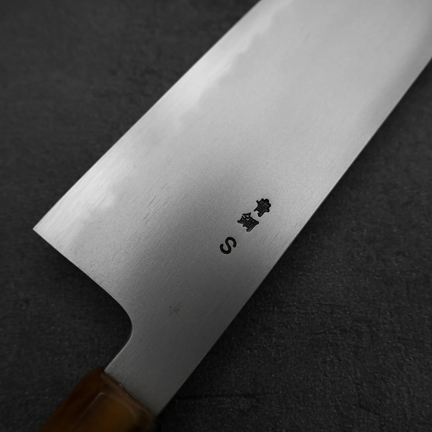A close-up of the Yoshikazu Tanaka AS tall gyuto 240mm shows its wooden handle and blade engraved with Japanese characters and an S, crafted by renowned blacksmith Yoshikazu Tanaka, resting on a dark textured background.