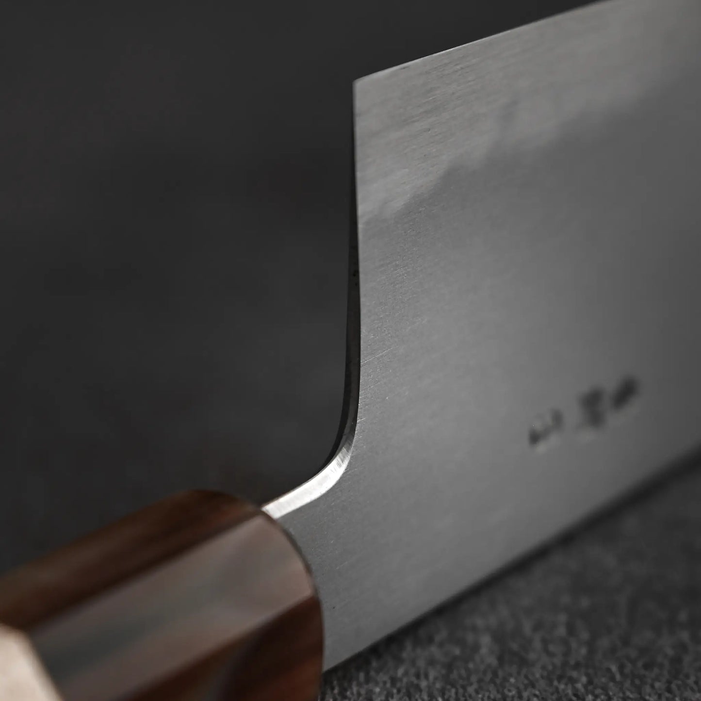 A close-up of the Yoshikazu Tanaka AS tall gyuto 240mm shows its sharp blade and fine wooden handle craftsmanship by renowned blacksmith Yoshikazu Tanaka, resting on a dark textured surface.