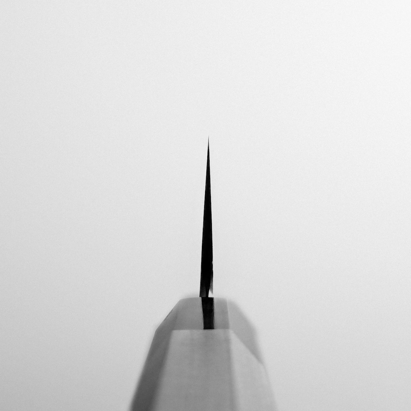 Close-up of the tip of a Yoshikazu Tanaka AS tall gyuto 210mm knife, photographed from the front against a plain, light background. The Yoshikazu Tanaka blades edge is centered and extends vertically.