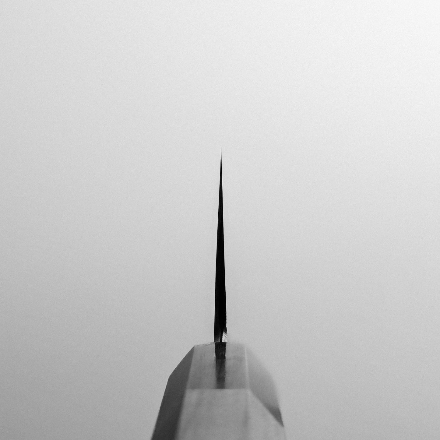 Close-up, centered view of the sharp tip of the Yoshikazu Tanaka AS tall gyuto 240mm chefs knife, with its symmetrical blade pointed at the viewer against a plain, light background.