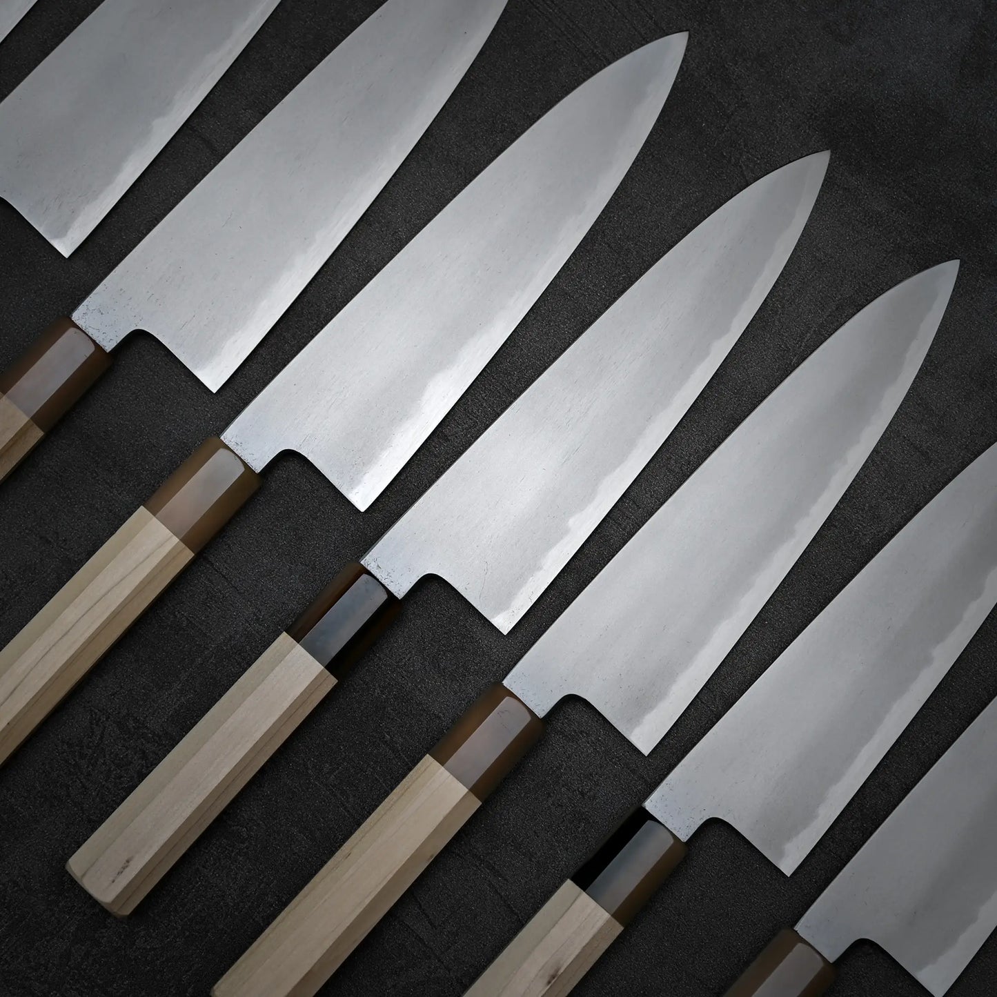 A row of kitchen knives, including the Yoshikazu Tanaka AS tall gyuto 210mm with a wooden handle and shiny blade, are arranged diagonally on a dark textured surface.