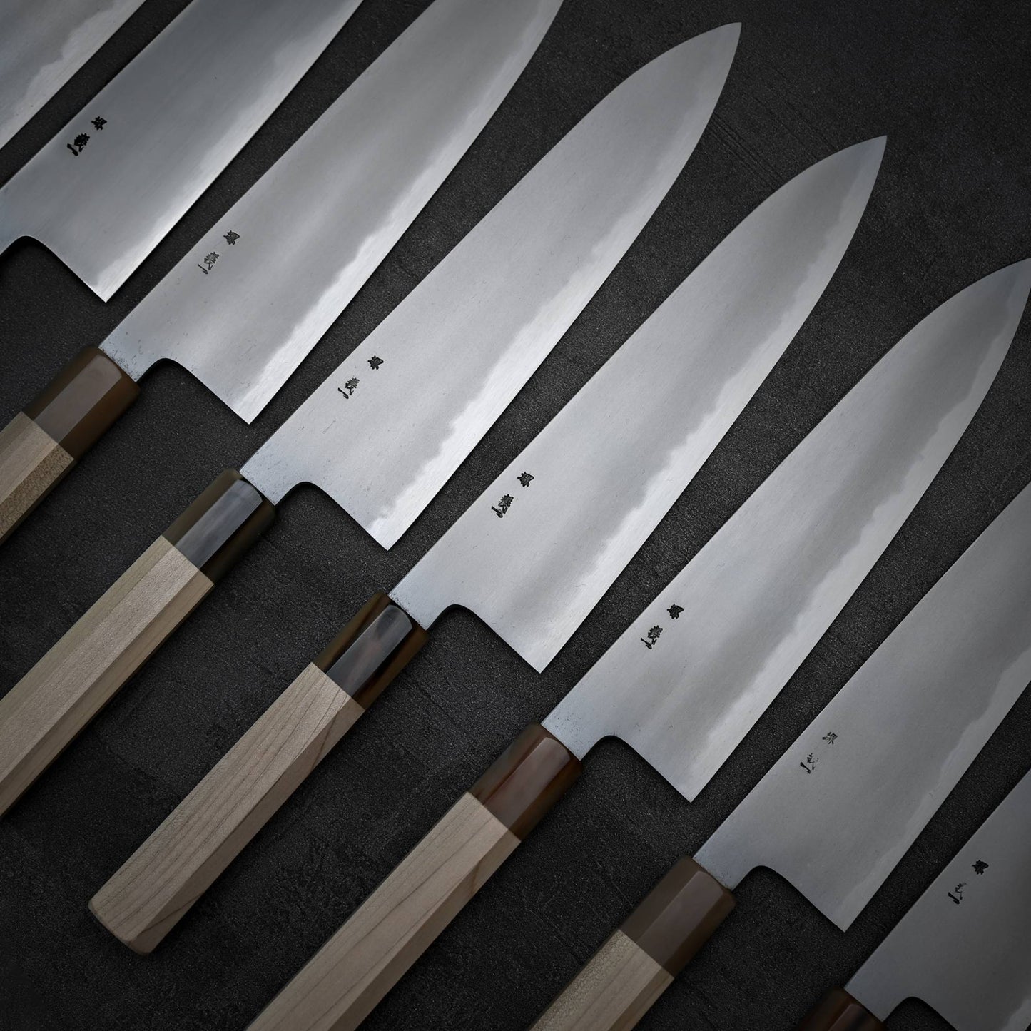 A row of Yoshikazu Tanaka AS tall gyuto 240mm knives, featuring wooden handles and shiny engraved blades, are arranged on a dark surface with the blades pointing to the right.