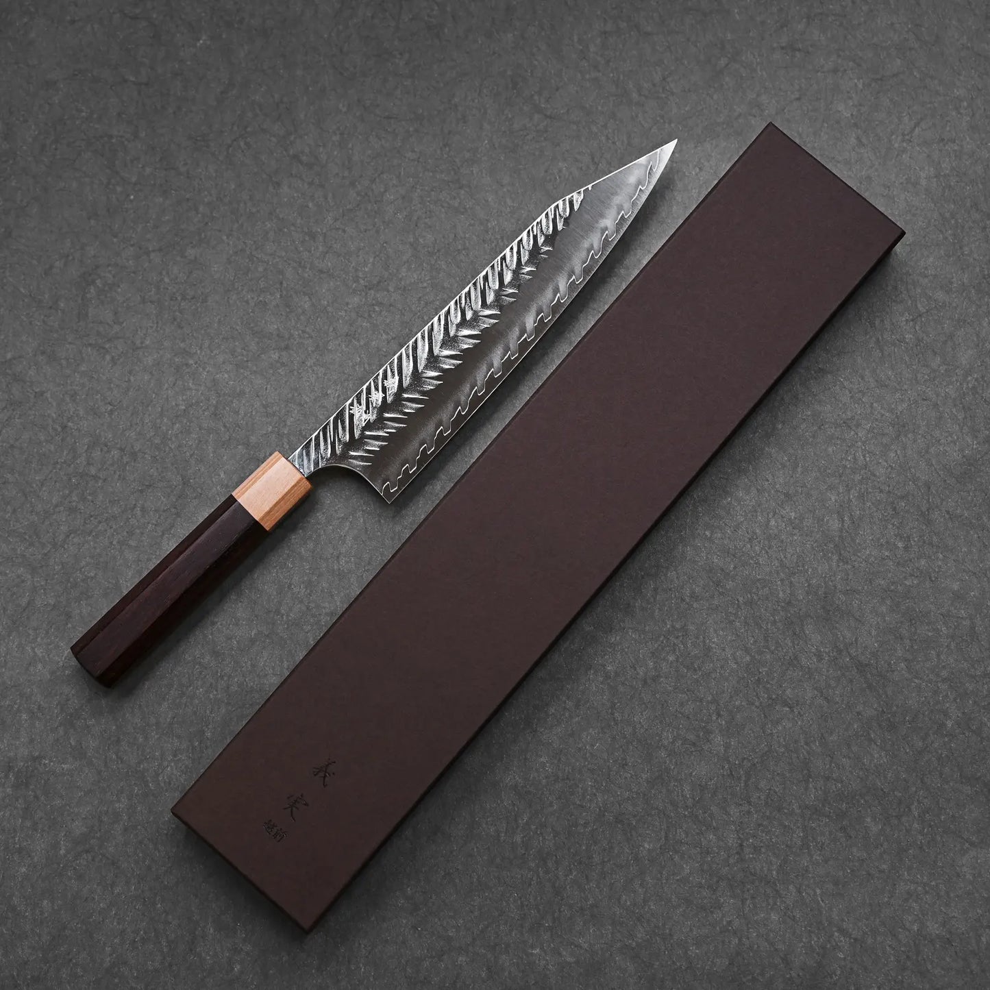 A Yoshimi Kato tsuchime STRIX kiritsuke gyuto 210mm chef’s knife with a patterned blade and wooden handle sits beside a long, brown SPG STRIX box on a dark textured surface.