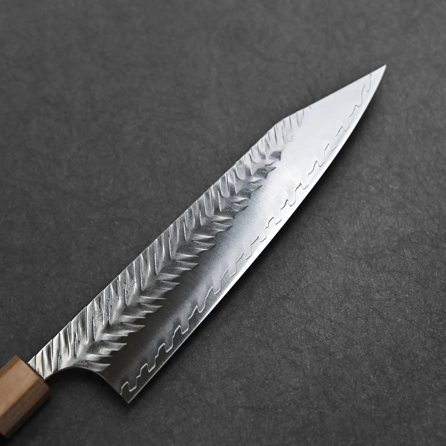 A Yoshimi Kato tsuchime STRIX kiritsuke gyuto 210mm kitchen knife by Yoshimi Kato with a wooden handle and a hammered, wavy-patterned blade rests on a dark, stone-like surface.