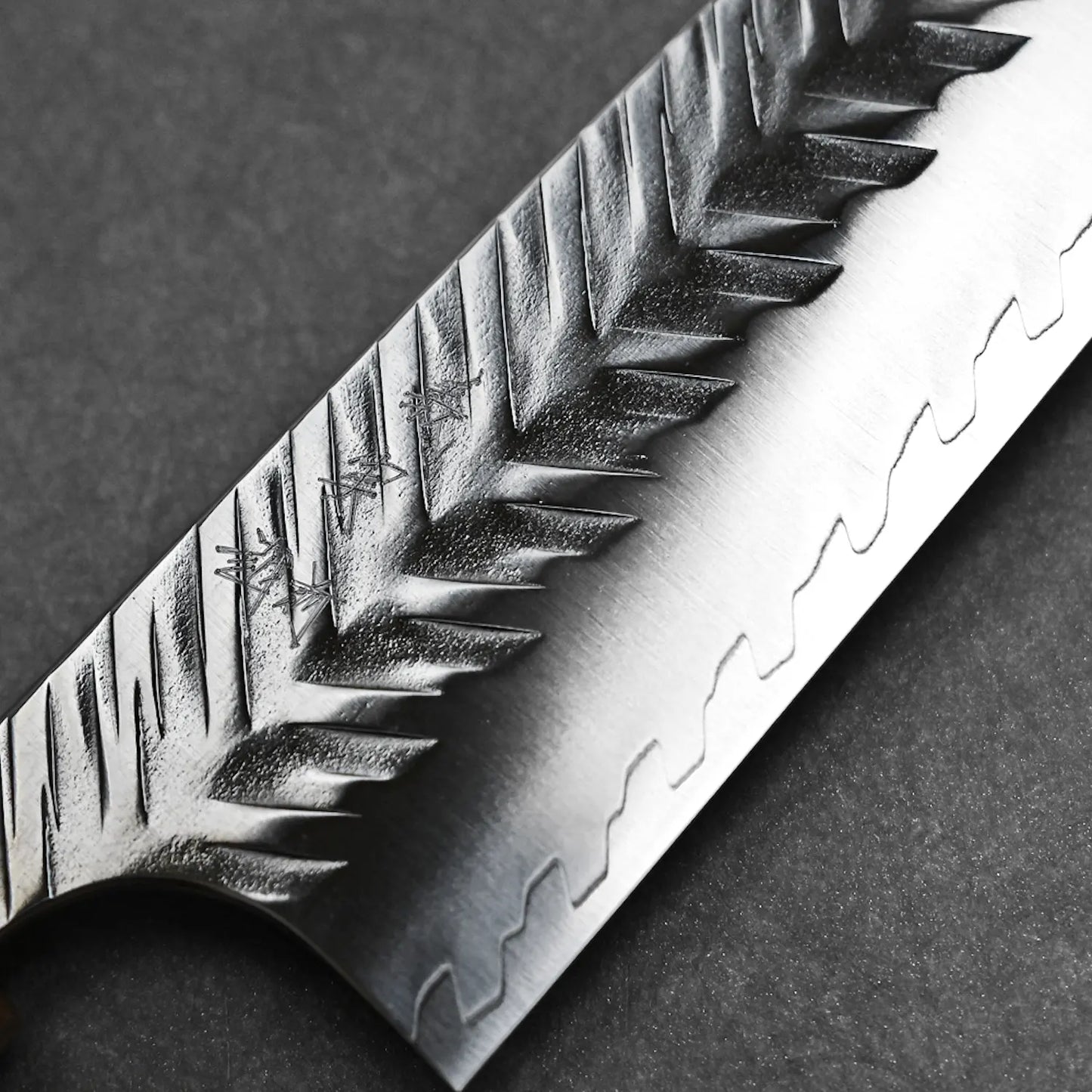 Close-up of a Yoshimi Kato tsuchime STRIX kiritsuke gyuto 210mm chef knife blade with a tree-branch-like etched pattern and jagged lower edge, displayed on a dark, textured surface. Brand: Yoshimi Kato.