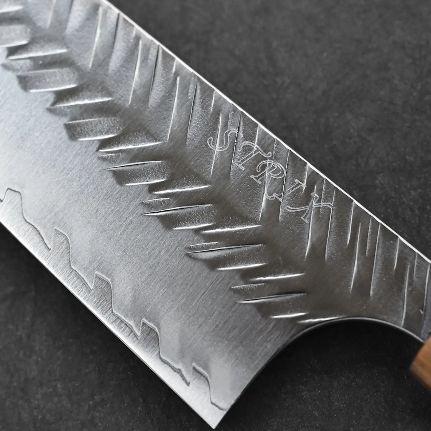 A close-up of the Yoshimi Kato tsuchime STRIX kiritsuke gyuto 210mm shows its jagged, textured blade with engraved letters. Crafted by Yoshimi Kato, the chef’s knife features a wooden handle and rests on a dark surface.