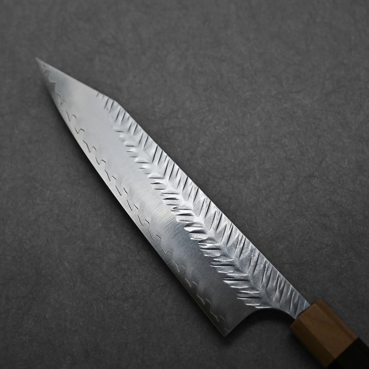 A close-up of the Yoshimi Kato tsuchime STRIX kiritsuke gyuto 210mm chef’s knife, featuring a patterned textured blade and wooden handle, on a dark gray surface.