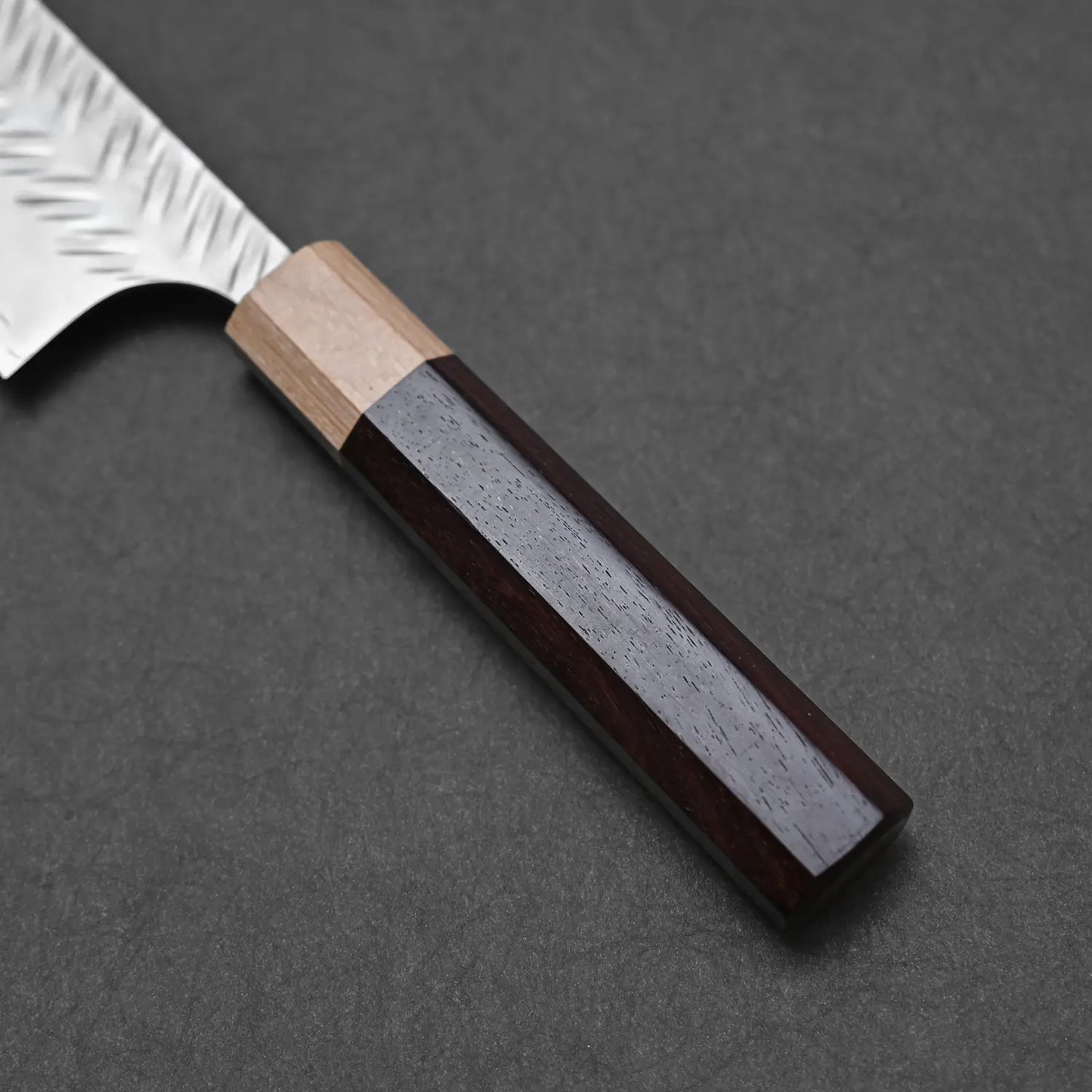 Close-up of the Yoshimi Kato tsuchime STRIX kiritsuke gyuto 210mm handle, featuring a dark wood lower and lighter wood upper section, with part of its textured blade visible against a dark, textured background.