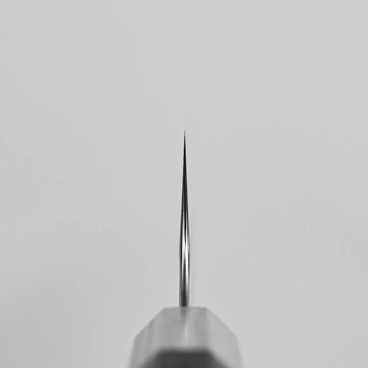 Close-up, centered view of the pointed tip of the Yoshimi Kato tsuchime STRIX kiritsuke gyuto 210mm knife against a light gray background. The metallic finish by Yoshimi Kato highlights its precision, with the sharp tip in clear focus.