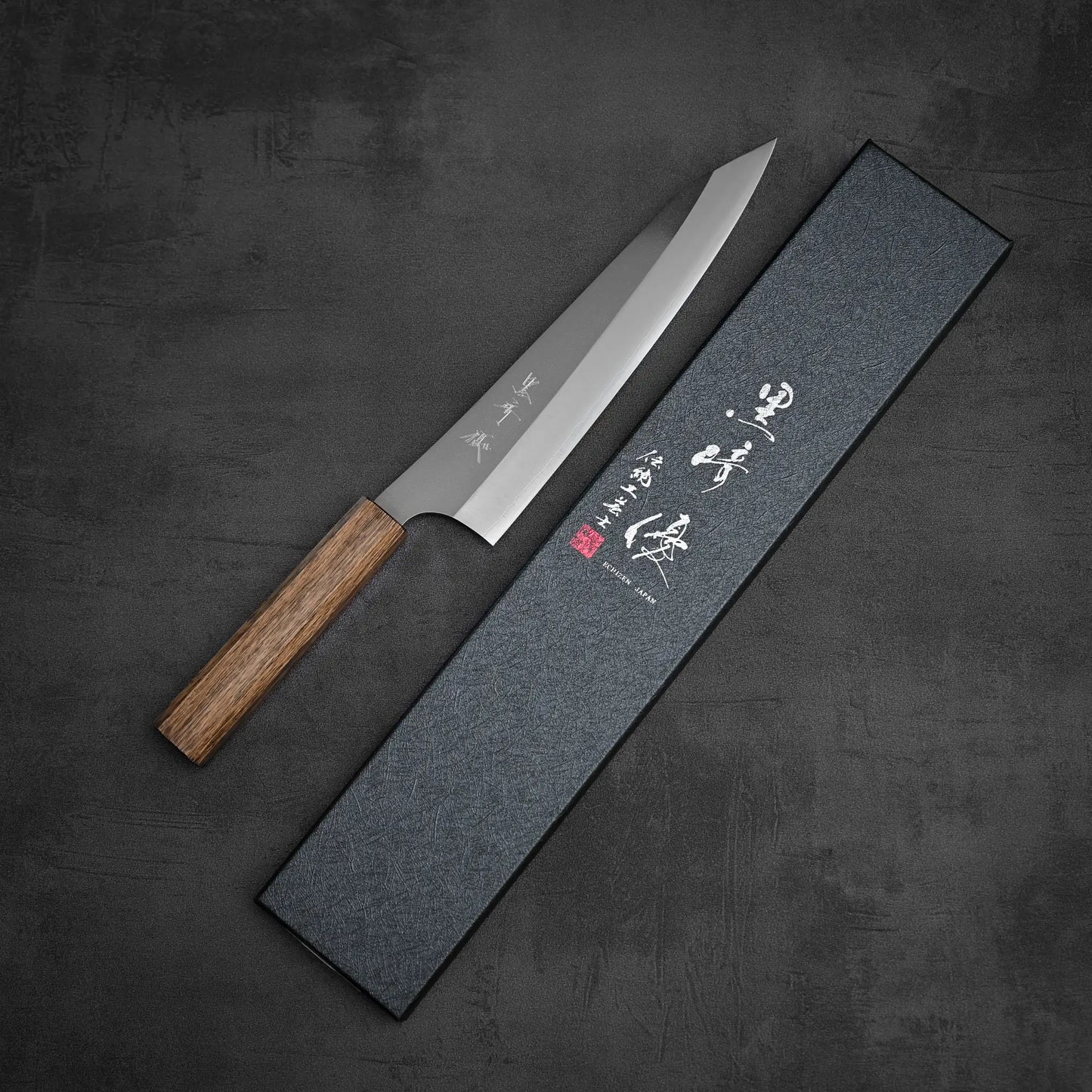 The Yu Kurosaki Gekko VG-XEOS kiritsuke gyuto 210mm, crafted by Yu Kurosaki, rests diagonally next to a textured box with Japanese characters on the lid, all set on a dark gray surface.