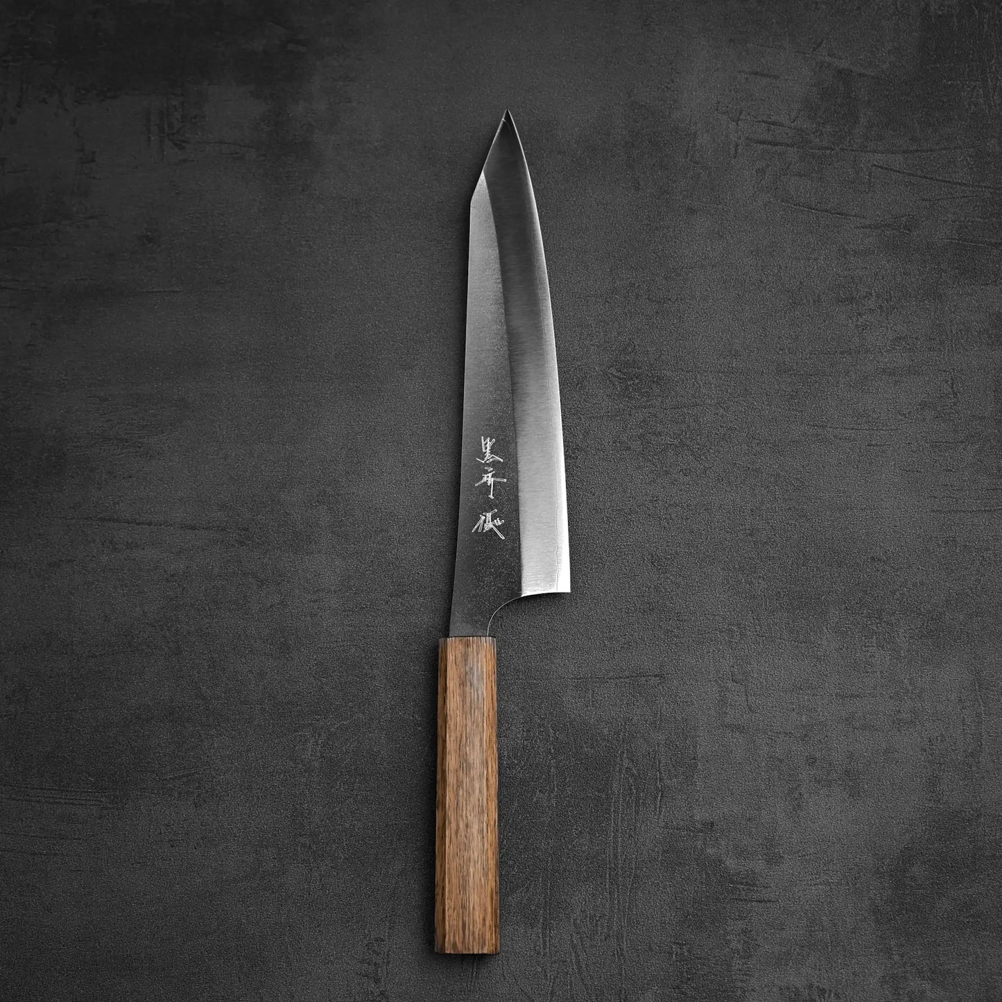 The Yu Kurosaki Gekko VG-XEOS kiritsuke gyuto 210mm chefs knife stands upright on a dark textured surface, showcasing its wooden handle and sharp, pointed VG-XEOS steel blade engraved with Japanese characters.
