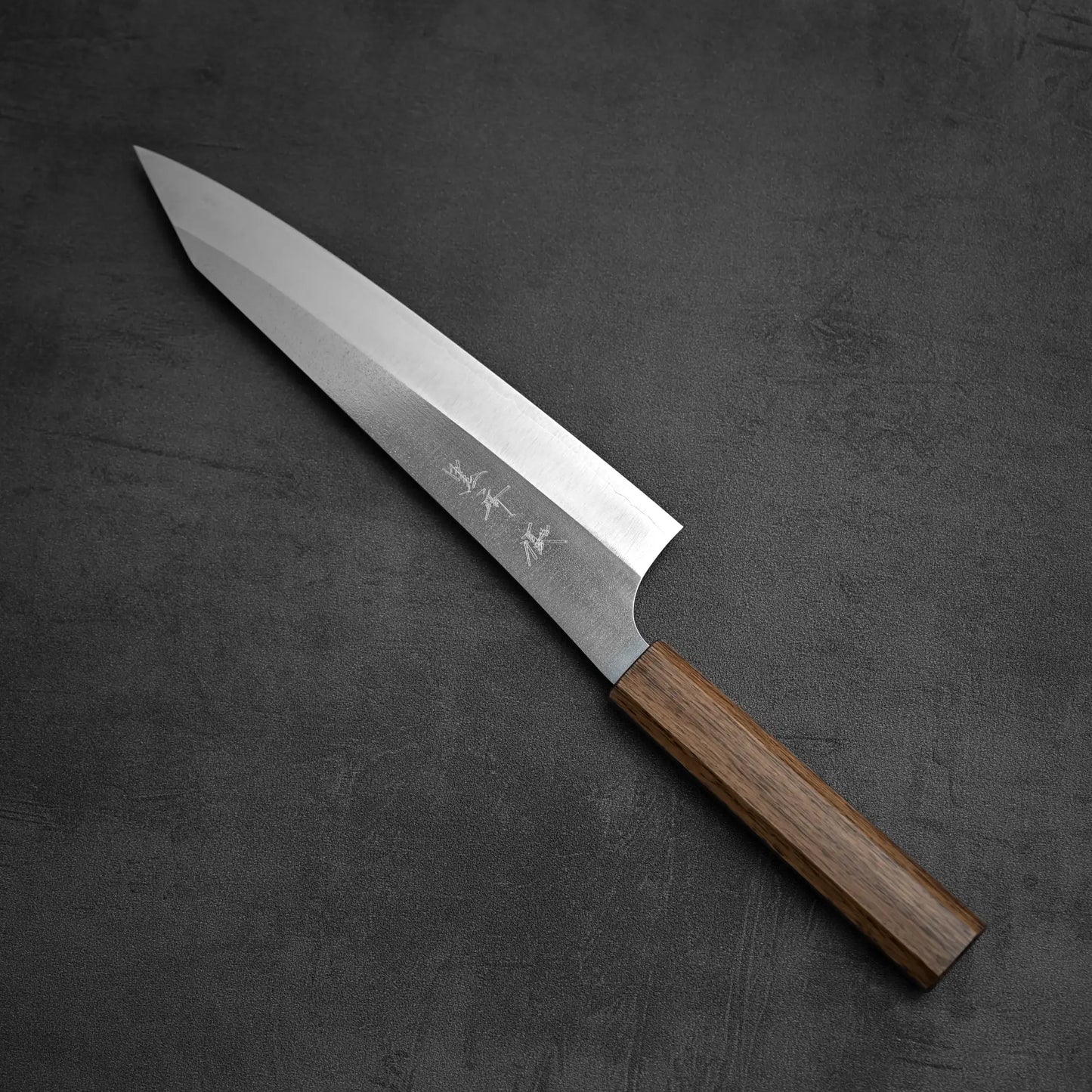 The Yu Kurosaki Gekko VG-XEOS kiritsuke gyuto 210mm, a Japanese chef’s knife with a silver blade etched with characters and a wooden handle, is displayed diagonally on a dark textured surface.