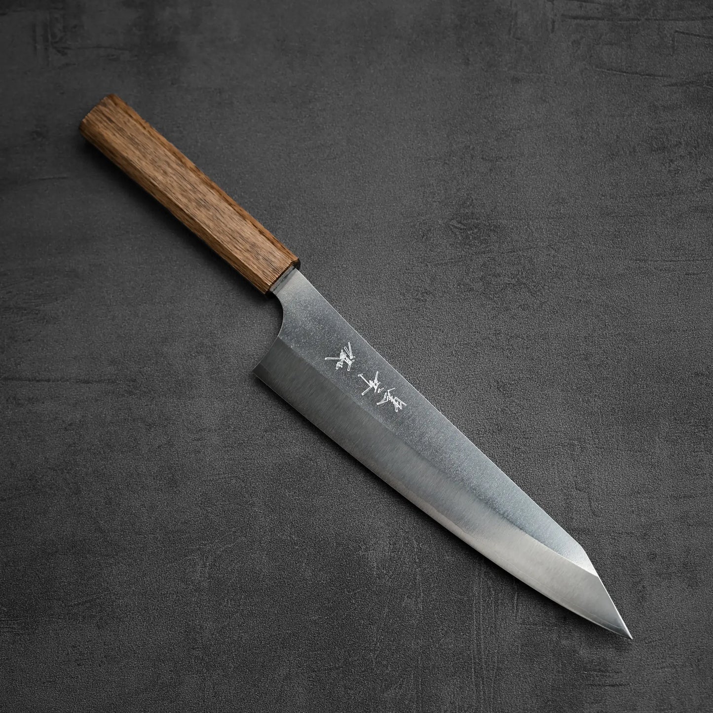 The Yu Kurosaki Gekko VG-XEOS kiritsuke gyuto 210mm, a Japanese-style chefs knife with a wooden handle and engraved kanji on its sharp blade, is shown resting on a dark textured surface.