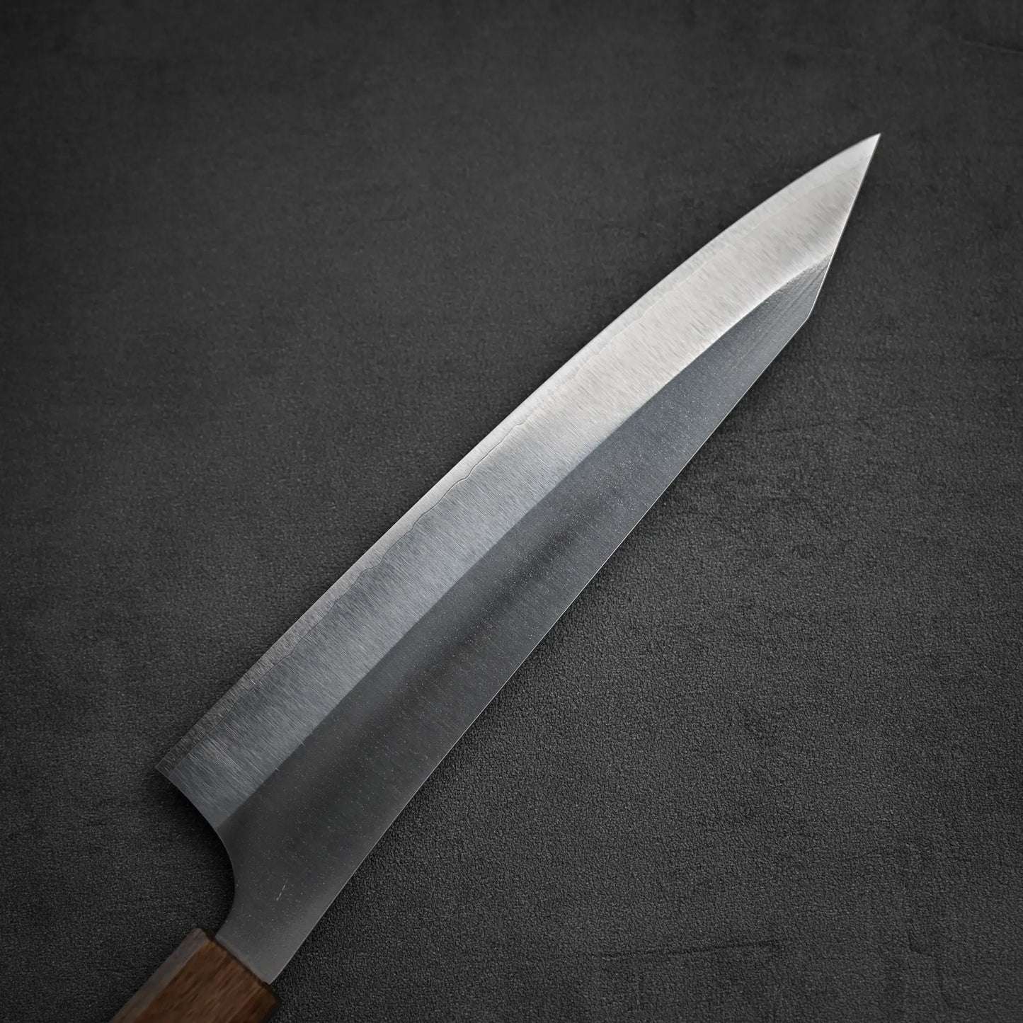 A close-up of the Yu Kurosaki Gekko VG-XEOS kiritsuke gyuto 210mm knife features its sharp VG-XEOS steel blade and wooden handle, resting diagonally on a dark, textured surface to highlight its clean edge and smooth finish.