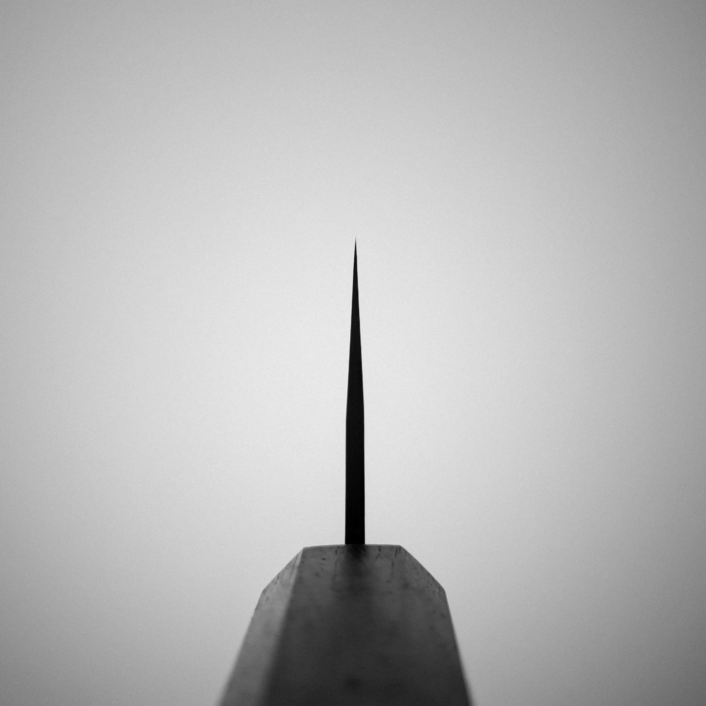 A black and white image shows the tip of a Yu Kurosaki Gekko VG-XEOS kiritsuke gyuto 210mm in sharp focus, centered against a plain light gray background. The narrow VG-XEOS steel point is clear, while the base is blurred in the foreground.