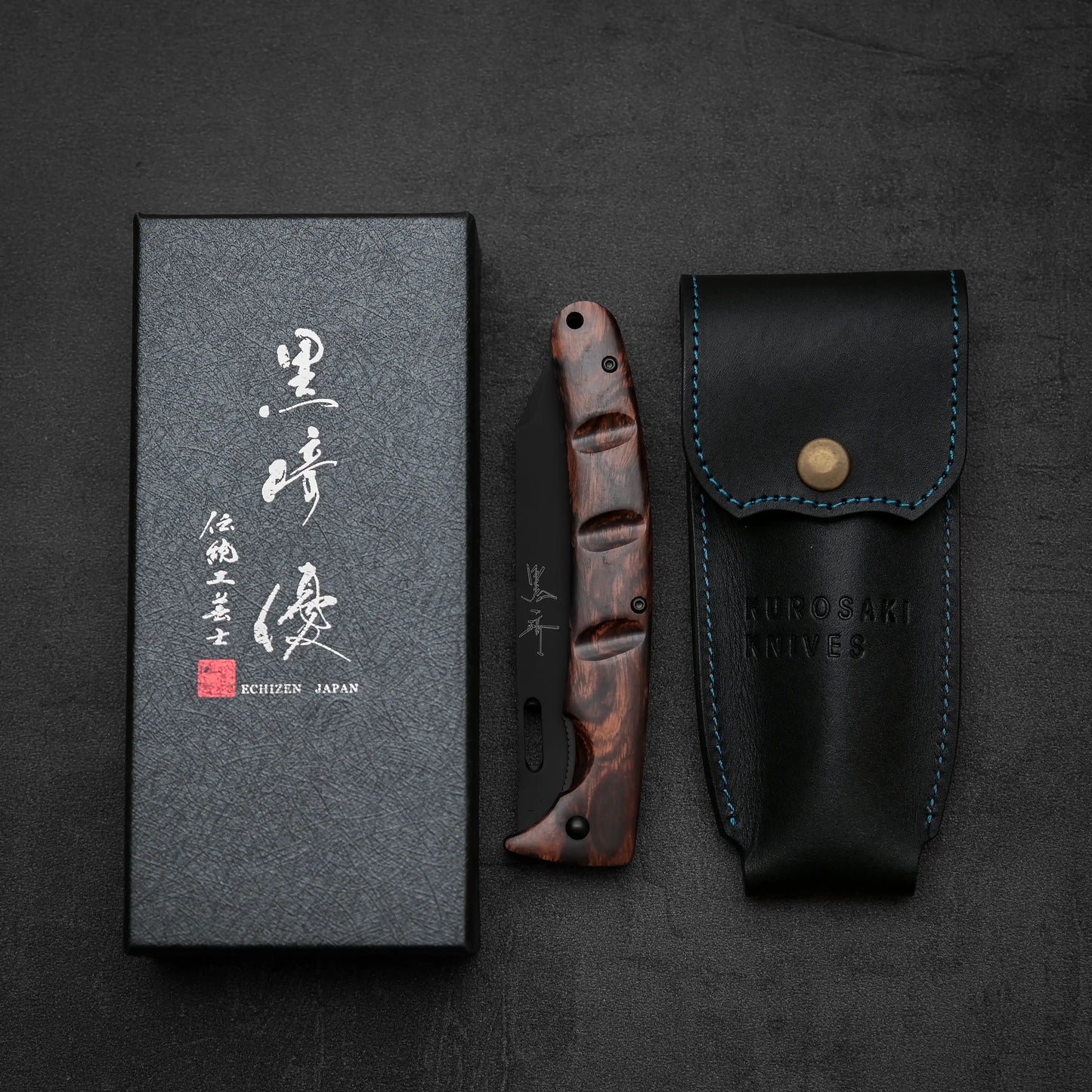 The Yu Kurosaki SG2 folding pocket knife with an ironwood handle is displayed between a black leather sheath with blue stitching and a black box labeled Yu Kurosaki Echizen Japan on a dark background.