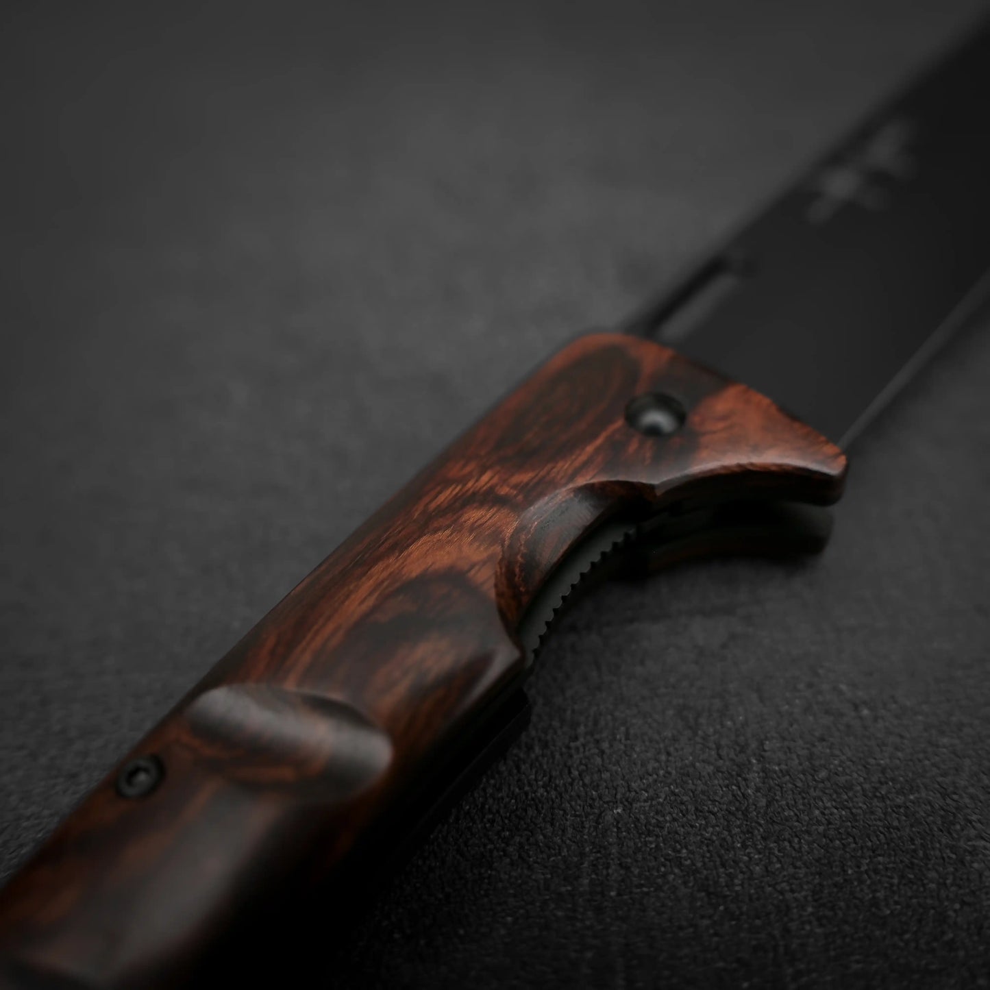 A close-up of the Yu Kurosaki SG2 folding pocket knife - ironwood by Yu Kurosaki, featuring a dark ironwood handle and partially visible black SG2 stainless steel blade on a textured dark surface.