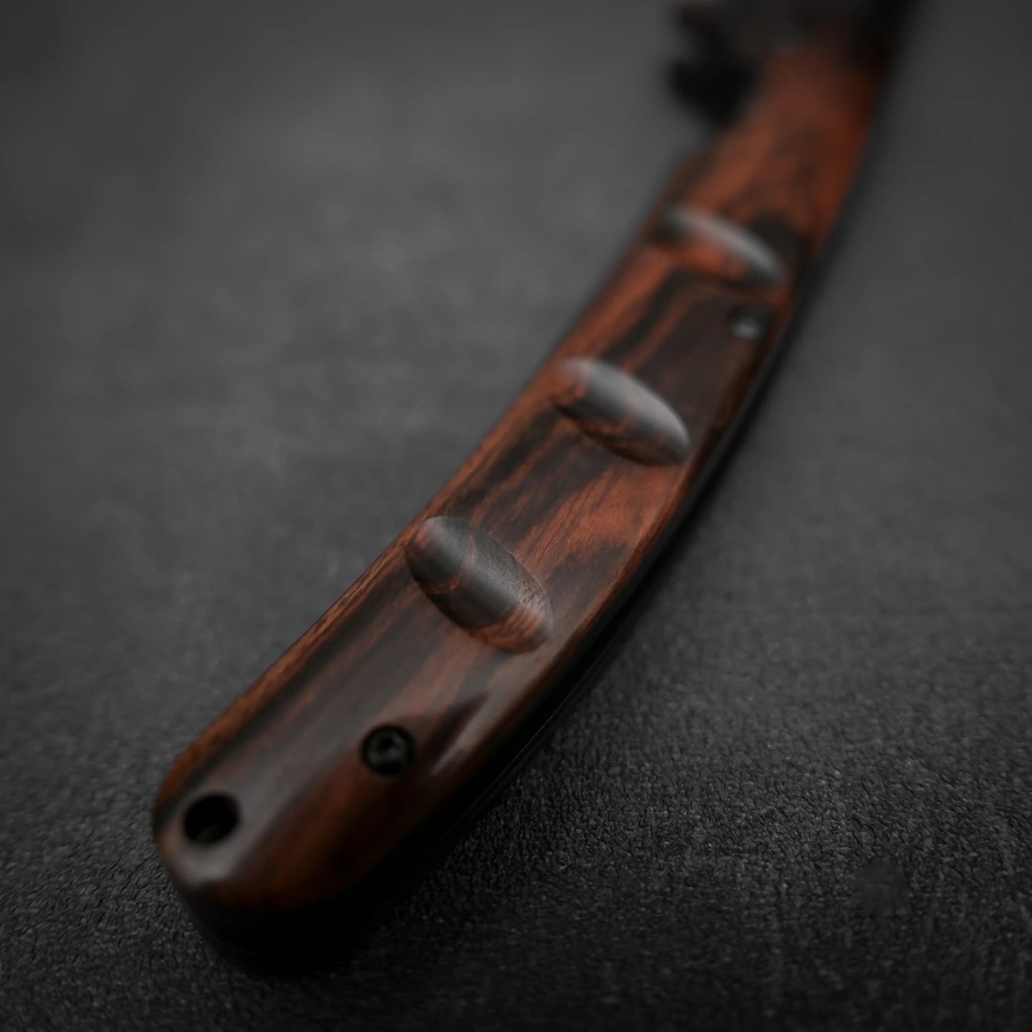 Close-up of the grooved dark ironwood handle with visible screws of the Yu Kurosaki SG2 folding pocket knife, resting on a textured black surface.