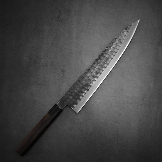 The Hinoura tsuchime kurouchi AS (stainless clad) gyuto 270mm by Hinoura, featuring a hammered blade and dark wooden handle, lies elegantly on a textured gray surface.