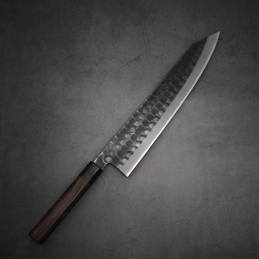 The Hinoura tsuchime kurouchi AS (stainless clad) kiritsuke gyuto 270mm by Hinoura, featuring a hammered Aogami Super steel blade and Macassar Ebony handle, rests on a dark gray textured surface.