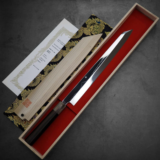 The Kenji Togashi shirogami#1 honyaki kiritsuke yanagiba 330mm with a dark handle is shown in a wooden box lined with red fabric, alongside its lid, certificate, and patterned sheath on a gray surface.