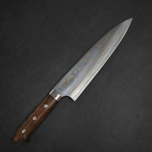 The Takeshi Saji rainbow damascus aogami#2 gyuto 210mm, featuring a dark wooden handle with three metal rivets and a striking Damascus blade, rests elegantly on a textured dark surface.