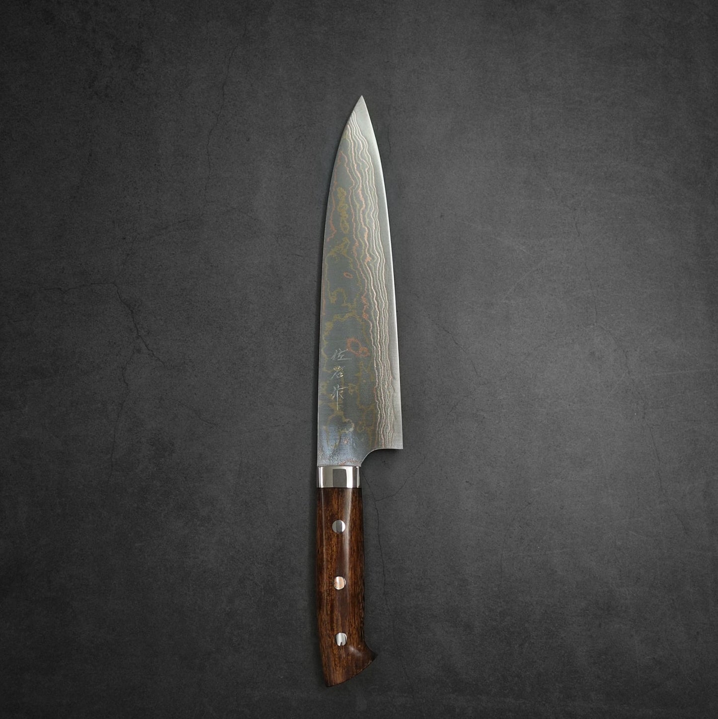The Takeshi Saji rainbow damascus aogami#2 gyuto 210mm, featuring a damascus pattern blade and wooden handle with three rivets, is placed on a dark, textured surface.