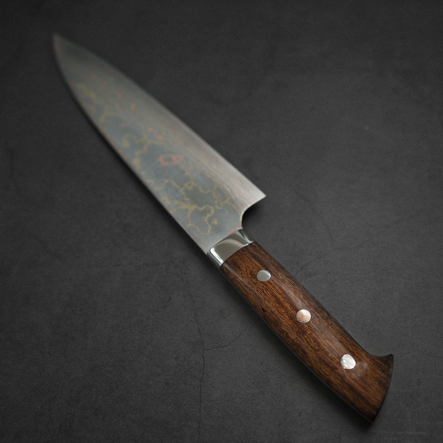 The Takeshi Saji rainbow damascus aogami#2 gyuto 210mm features a striking damascus steel blade and a wooden handle with three metal rivets, displayed on a dark, textured surface.