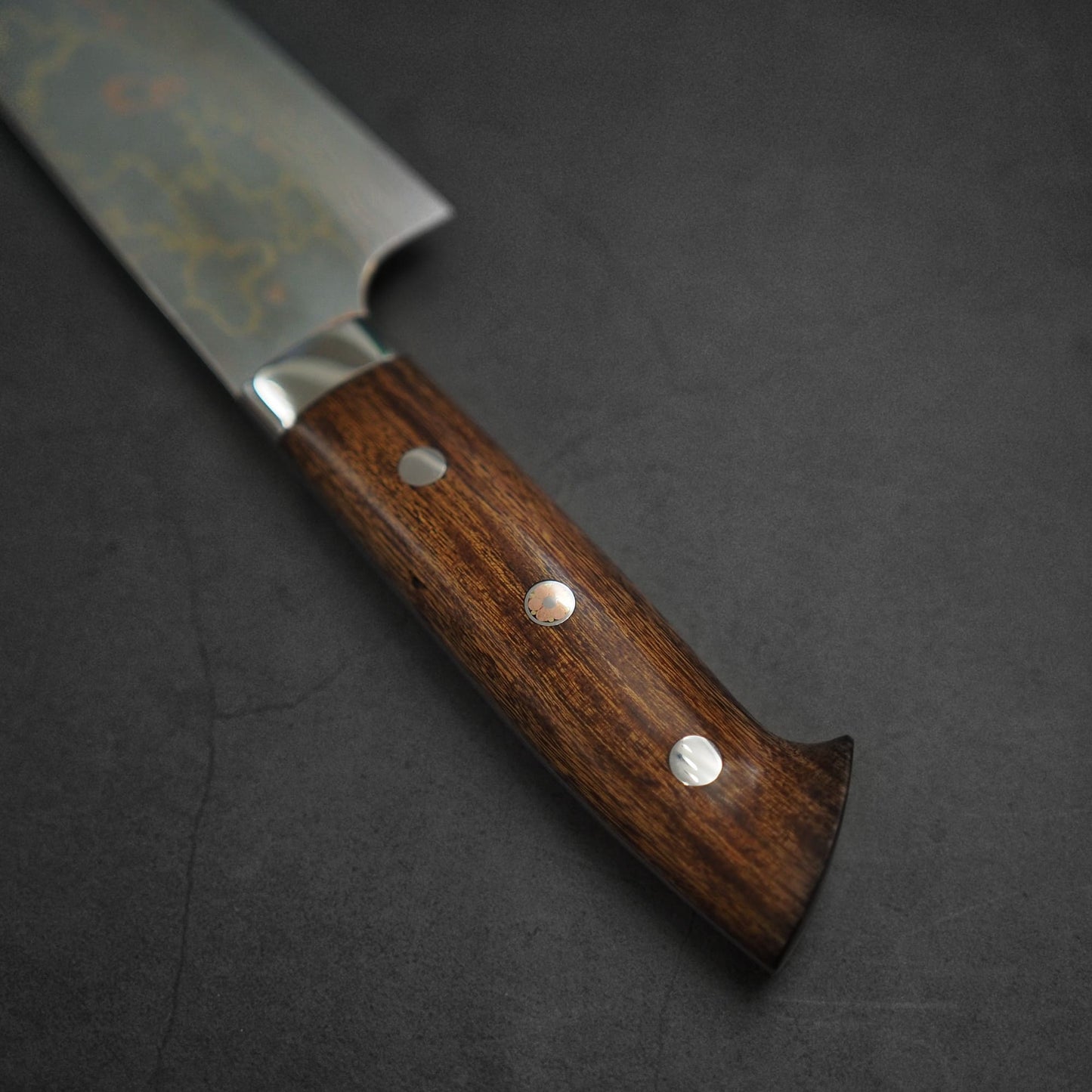 Close-up of the Takeshi Saji rainbow damascus aogami#2 gyuto 210mms polished dark wood handle with three metal rivets, set against a dark textured background. The blade features a subtle damascus pattern, partially visible at the top left.
