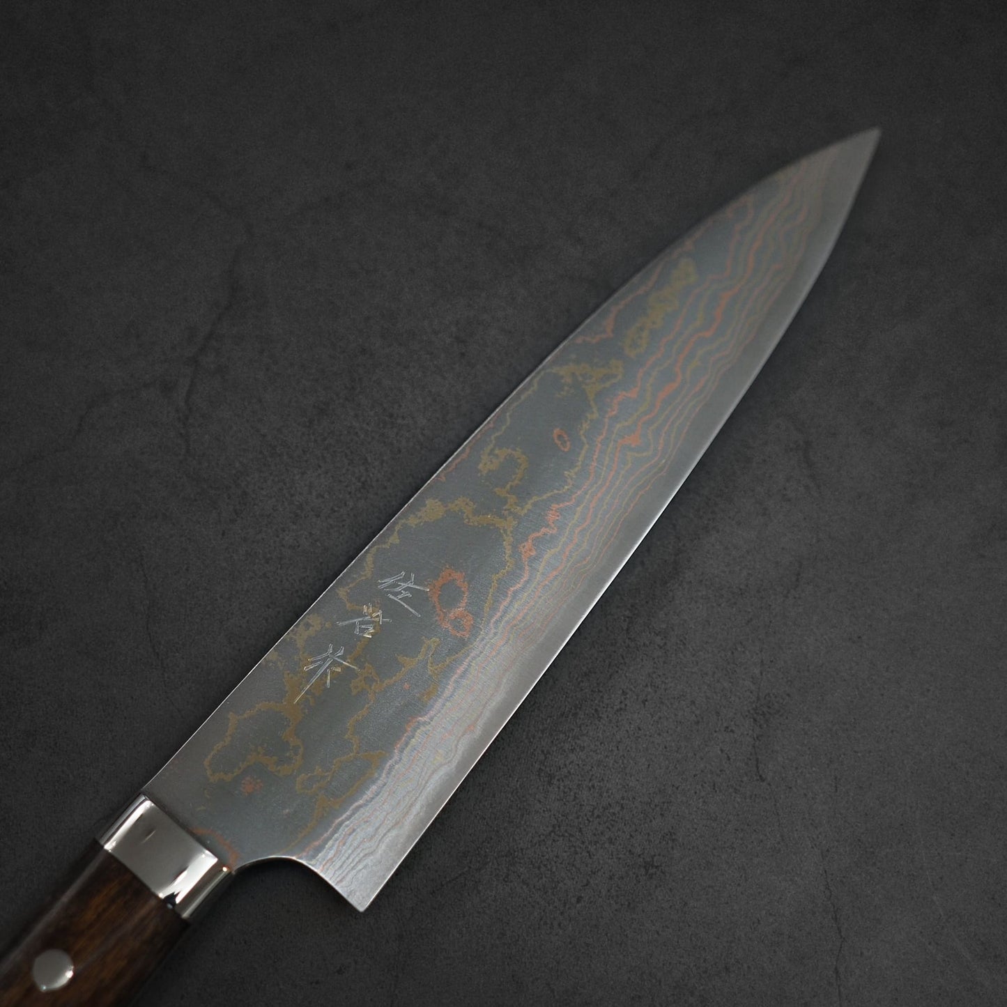 The Takeshi Saji rainbow damascus aogami#2 gyuto 210mm features a dark wooden handle and a striking damascus-pattern blade, elegantly displayed on a dark stone surface.