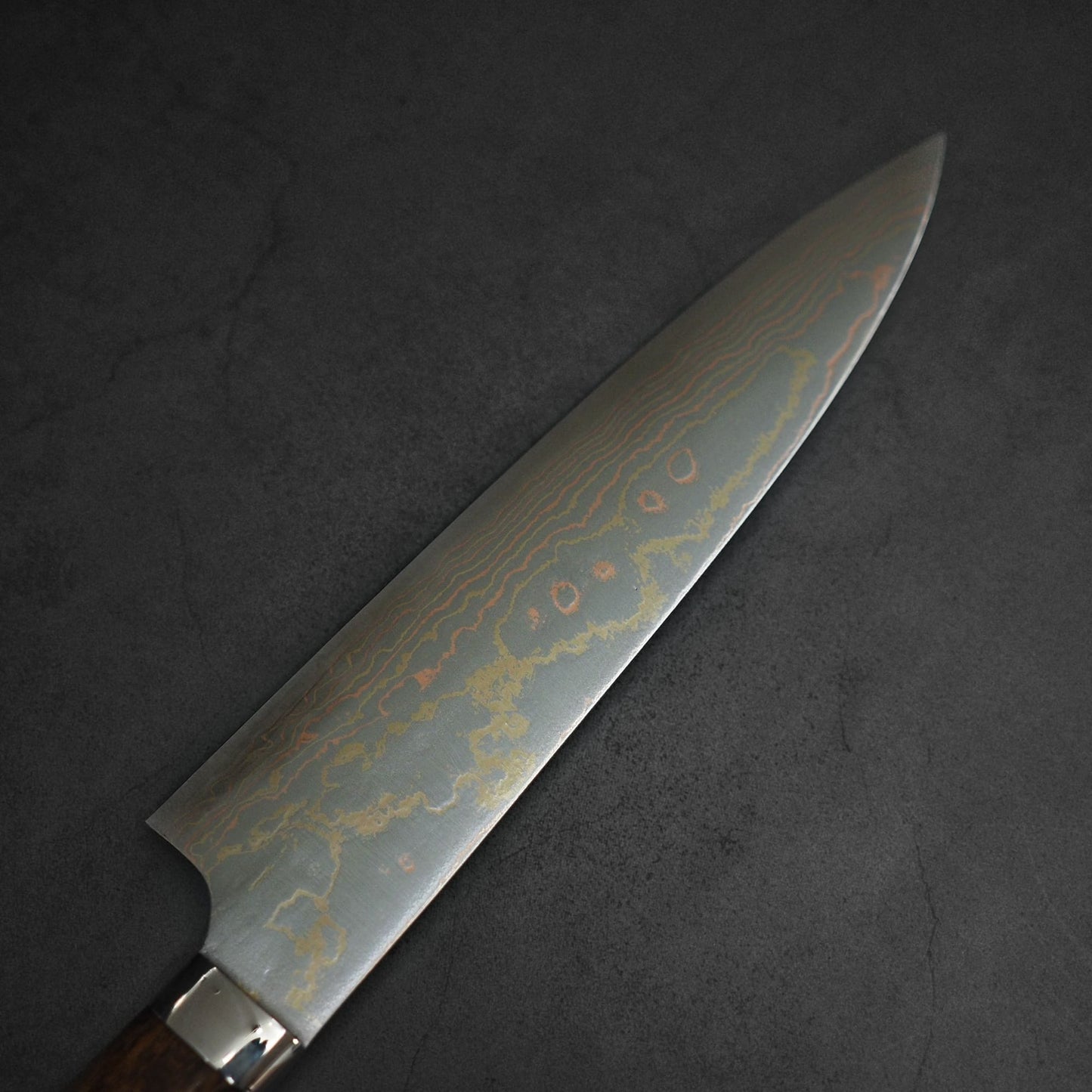 A close-up of the Takeshi Saji Rainbow Damascus Aogami#2 Gyuto 210mm showcases its brands signature layered steel and intricate wavy damascus pattern, displayed on a dark, textured surface.