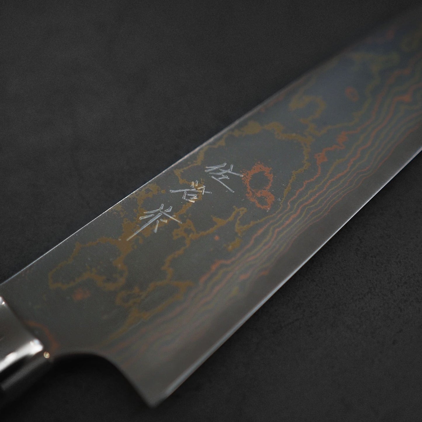 A close-up of the Takeshi Saji rainbow damascus aogami#2 gyuto 210mm blade, featuring a striking damascus pattern and kanji engraving, by Takeshi Saji, photographed against a dark background.