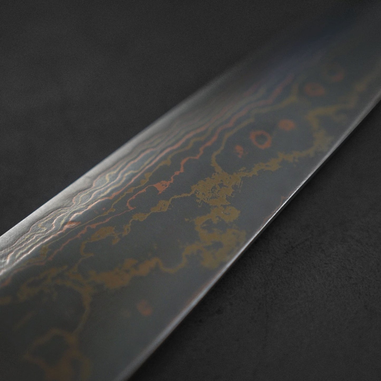 A close-up of the Takeshi Saji rainbow damascus aogami#2 gyuto 210mm showcases its intricate wavy damascus pattern on the blade, set against a dark, blurred background.