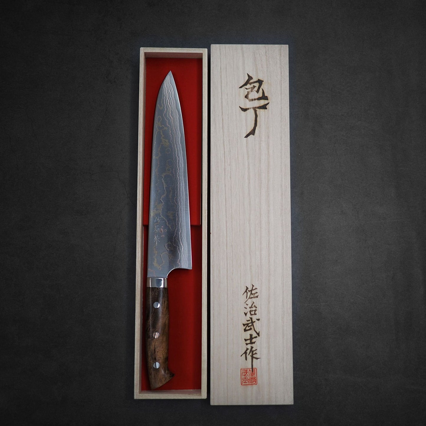 The Takeshi Saji rainbow damascus aogami#2 gyuto 240mm, featuring a patterned blade and wooden handle, is displayed in a wooden box with Japanese characters on the lid against a dark background.