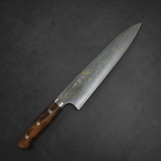 A Takeshi Saji rainbow damascus aogami#2 gyuto 240mm with a dark wooden handle lies diagonally on a textured dark surface.