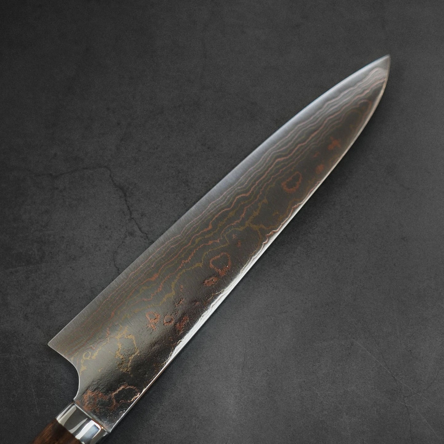 A close-up of the Takeshi Saji Rainbow Damascus Aogami#2 Gyuto 240mm chef’s knife, featuring a patterned blade and dark handle, resting on a textured dark gray surface.