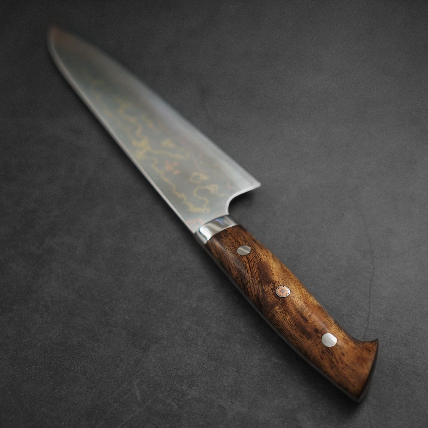 A Takeshi Saji rainbow damascus aogami#2 gyuto 240mm chef’s knife with a polished wooden handle rests on a dark textured surface.
