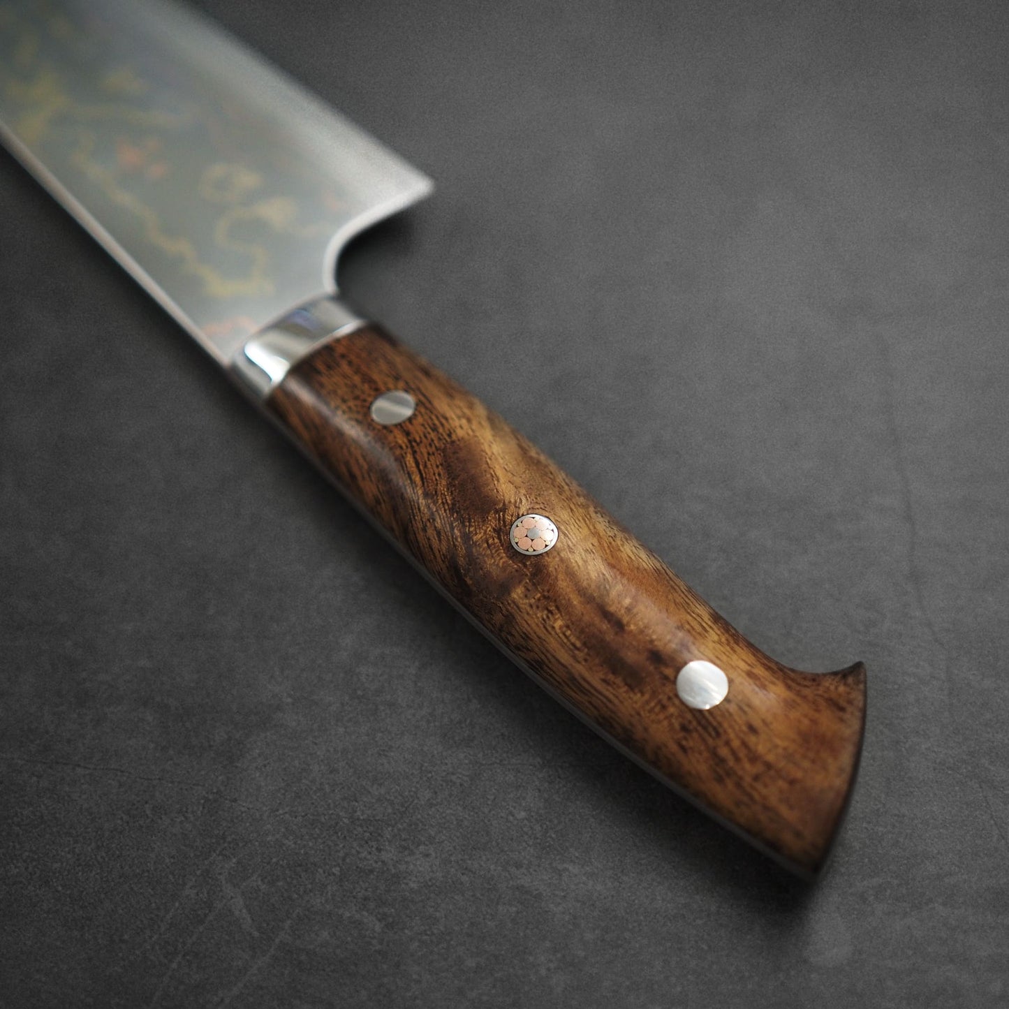 A close-up of the Takeshi Saji rainbow damascus aogami#2 gyuto 240mm chef’s knife handle, made from polished dark wood with three metal rivets, rests on a matte surface; its damascus aogami#2 steel blade appears at the top left.