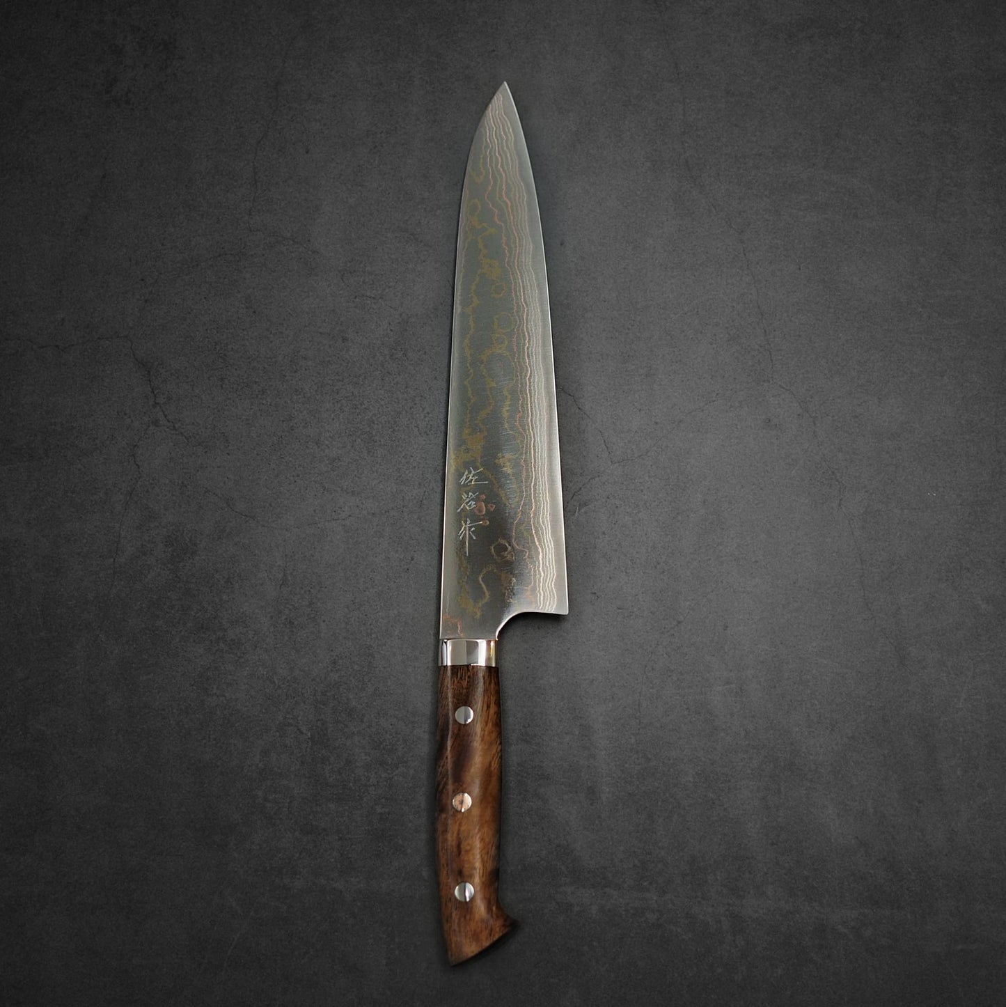 A Takeshi Saji rainbow damascus aogami#2 gyuto 240mm with a polished wooden handle rests on a dark, textured surface.