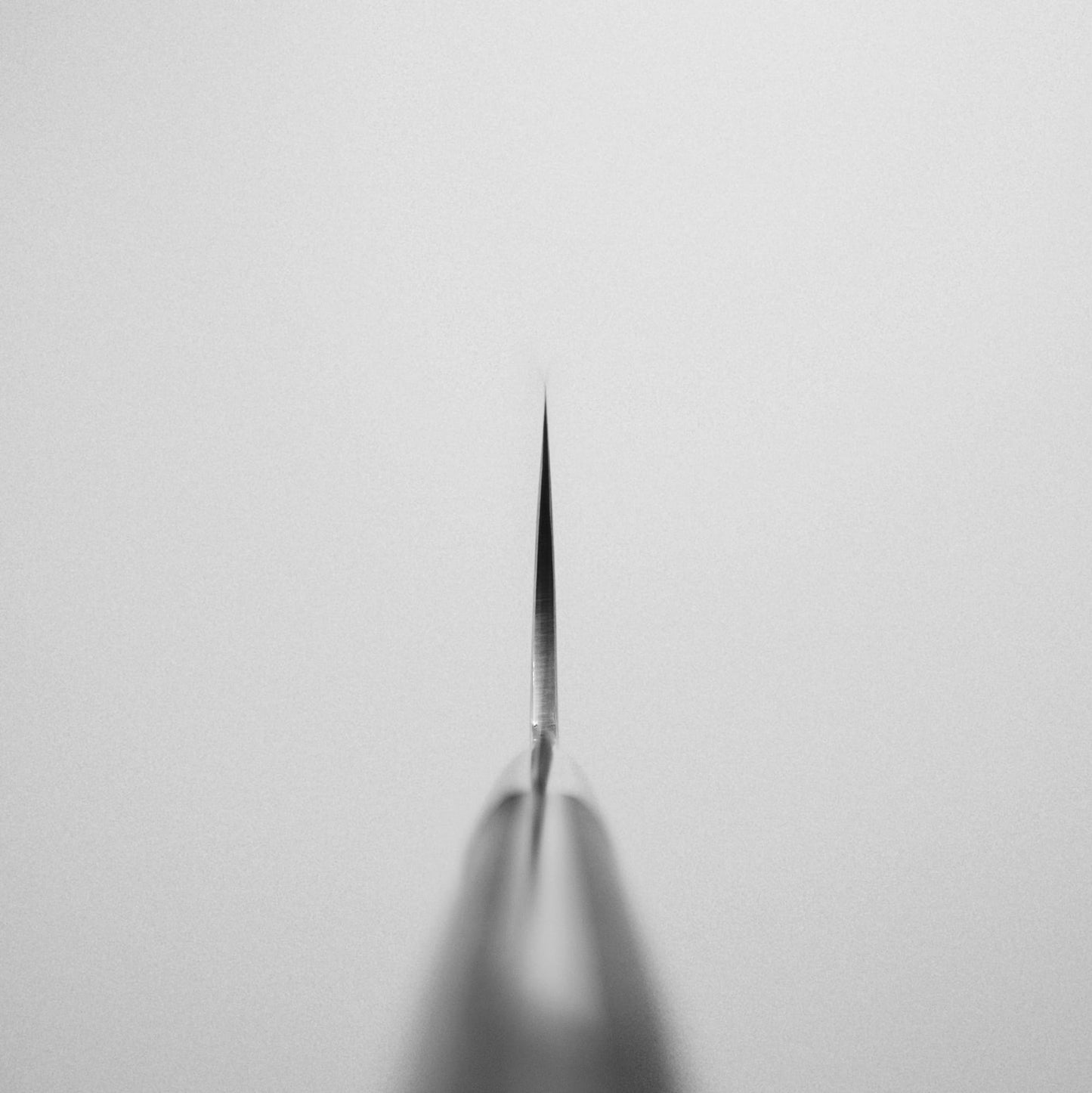 A close-up, centered view of the pointed tip of a Takeshi Saji rainbow damascus aogami#2 gyuto 240mm knife against a plain, light background. The image is black and white with a minimal composition.